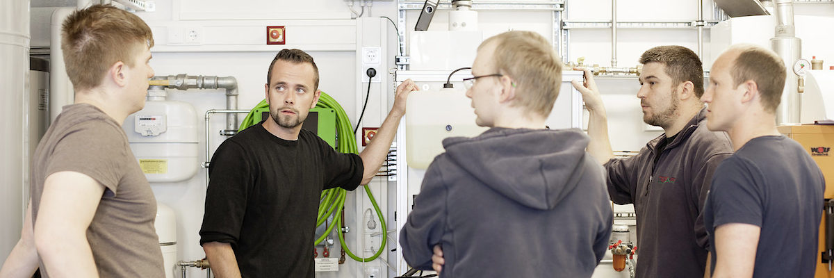 Die Handwerksjunioren wollen das Handwerk mitgestalten und dafür werben Fünf junge Männer stehen in einem Raum mit Geräten. Vier davon schauen den Mann ganz links an, der scheinbar etwas sagt.