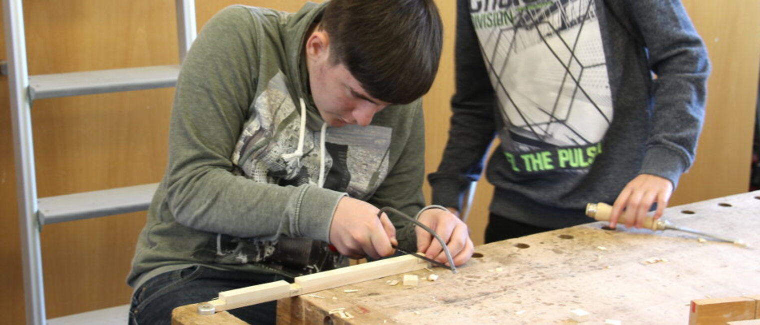 In der Lernwerkstatt lernen Schüler das Bauhandwerk kennen. Ein Jugendlicher mit dunklen Haaren sitzt am Tisch in einem Werkraum und sägt ein Stück aus einer Holzleiste.