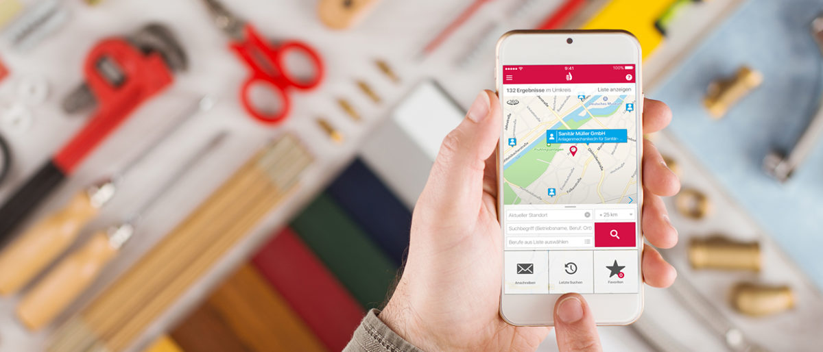 Mit der kostenfreien App Handwerkerradar können Verbraucher den passenden Handwerksbetrieb in ihrer Nähe finden. Eine Männerhand hält ein Smartphone. Auf dessen Display ist eine Karte zu sehen. Im Hintergrund sind verschwommen Werkzeuge zu sehen.