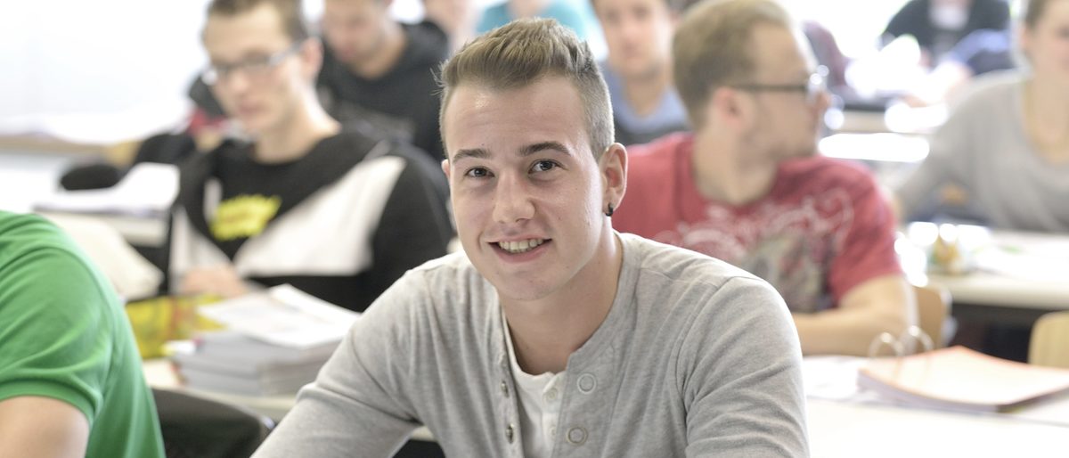 Das BTZ bietet auch individuelle Seminare für Firmen an. In einem Seminarraum sitzen junge Menschen an Tischen. Ein junger Mann in der Mitte lächelt in die Kamera.
