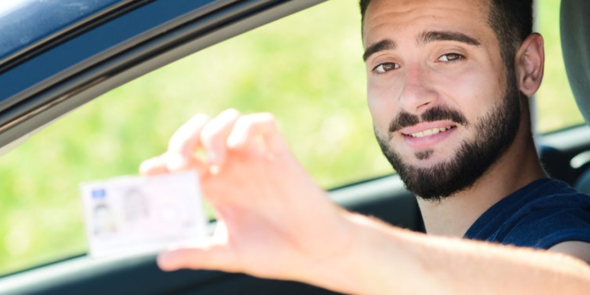 Ein junger Mann schaut aus dem geöffneten Fahrerfenster eines Pkws und hält seinen Führerschein in die Kamera.