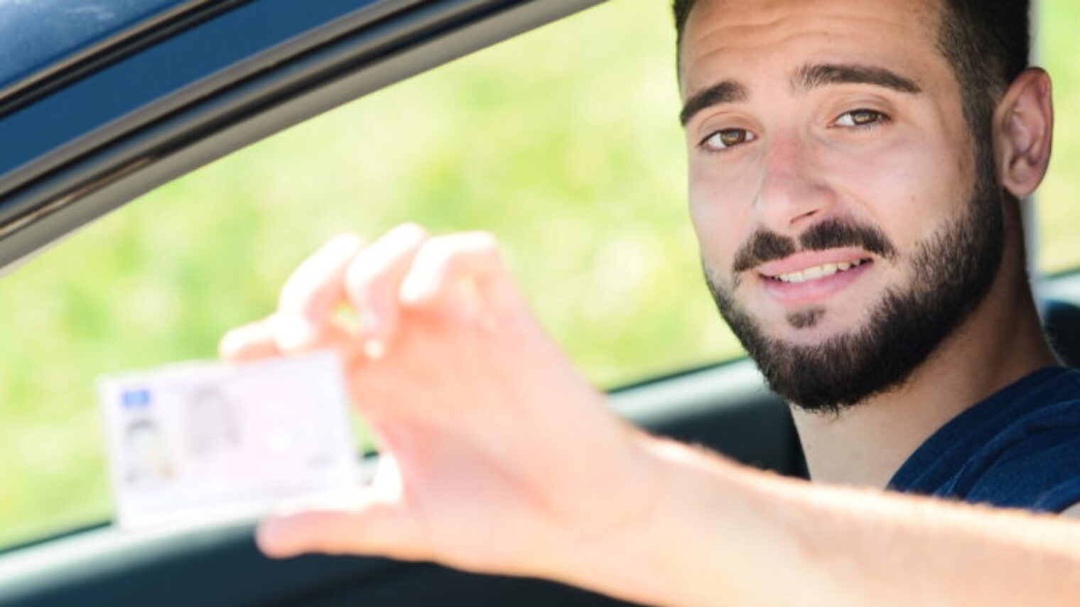 Ein junger Mann schaut aus dem geöffneten Fahrerfenster eines Pkws und hält seinen Führerschein in die Kamera.