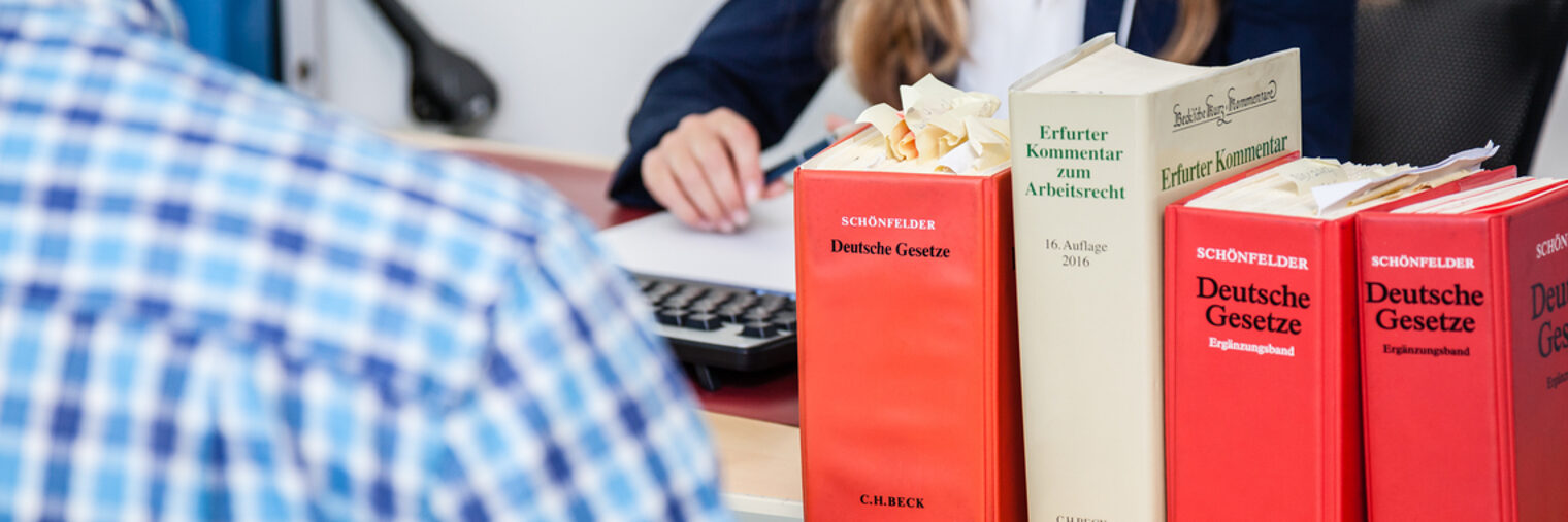 Vier dicke Gesetzesbücher stehen rechts vorn im Bild. Im Hintergrund sieht man eine Frauenhand, die einen Stift hält. 