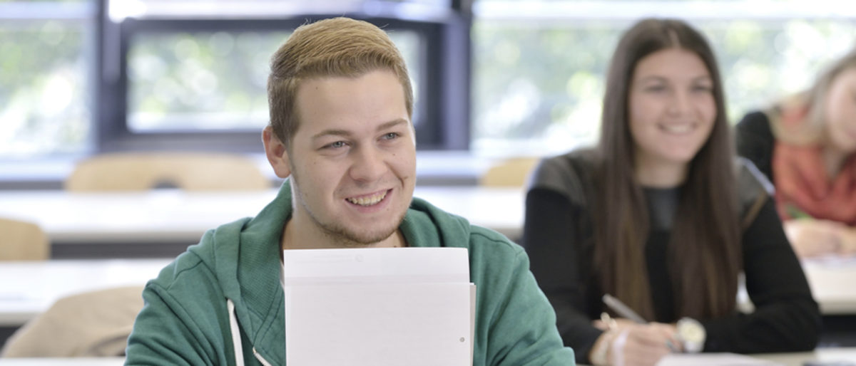 Lernen macht Spaß. Ein junger Mann lächelt in die Kamera. Er hält ein paar Blätter vor sich. Im Hintergrund zwei junge Frauen an einem Schultisch, die eine lächelt, die andere schreibt etwas.