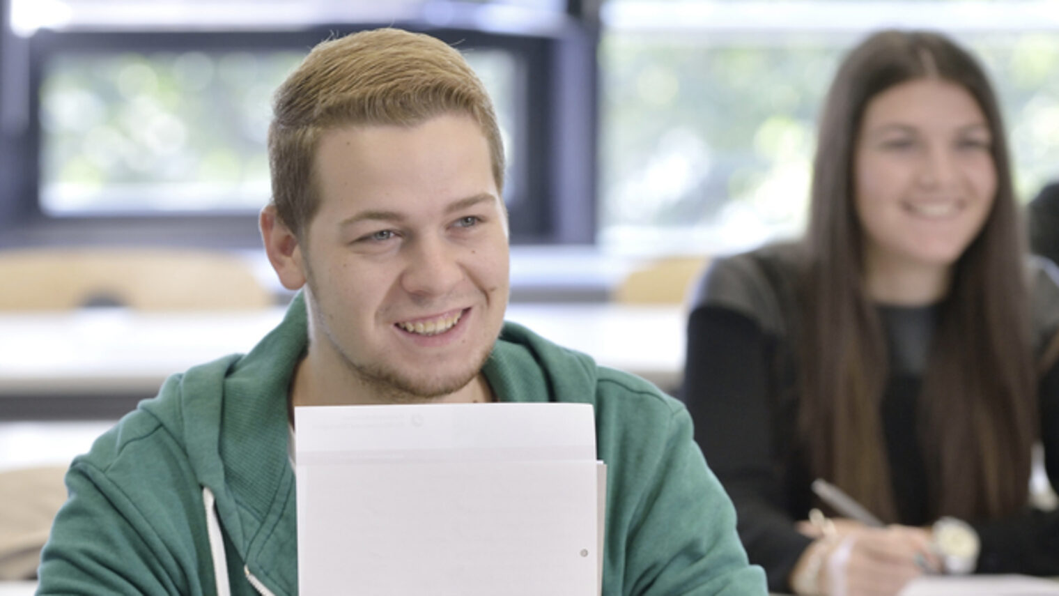 Ein junger Mann lächelt in die Kamera. Er hält ein paar Blätter vor sich. Im Hintergrund zwei junge Frauen an einem Schultisch, die eine lächelt, die andere schreibt etwas.