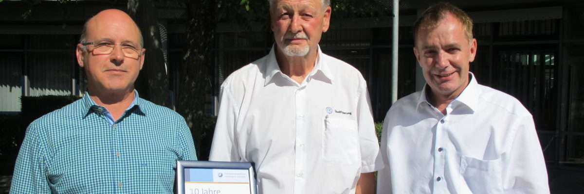 Für das ehrenamtliche Engagement gab es eine Urkunde für Dieter Fuchs (Mitte) und viel Lob von Wolfgang Lauckner (links), Vorsitzender des Prüfungsausschusses, und BTZ-Leiter Johannes Richter. Drei Männer sehen in die Kamera. Sie stehen vor einem Baum. Der Mann links hält eine gerahmte Urkunde in der Hand.