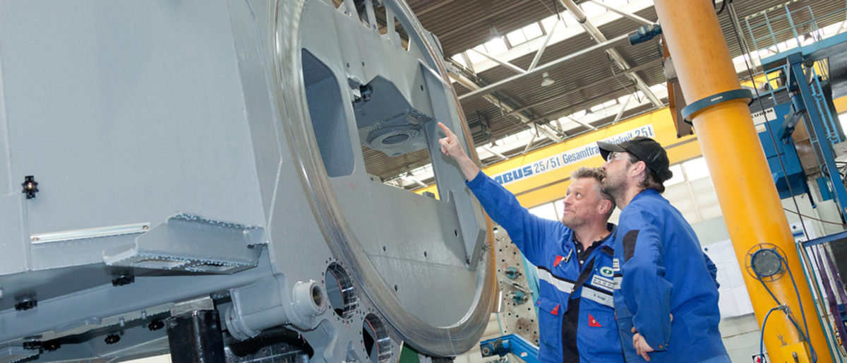 In den meisten Betrieben laufen die Maschinen derzeit auf Hochtouren. Zwei Männer in Arbeitskleidung stehen in einer Fabrikhalle und betrachten ein großes Teil aus Metall.