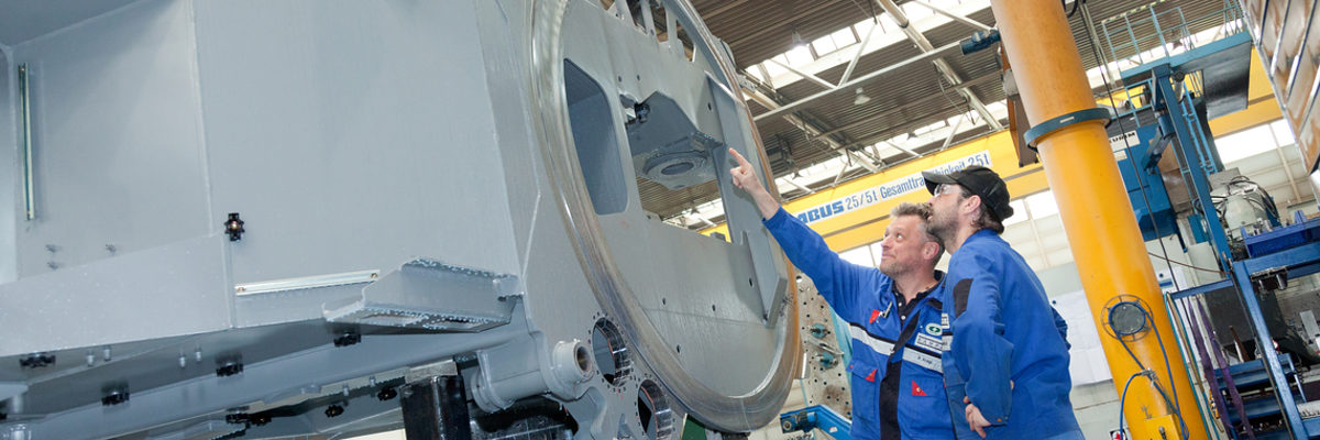In den meisten Betrieben laufen die Maschinen derzeit auf Hochtouren. Zwei Männer in Arbeitskleidung stehen in einer Fabrikhalle und betrachten ein großes Teil aus Metall.