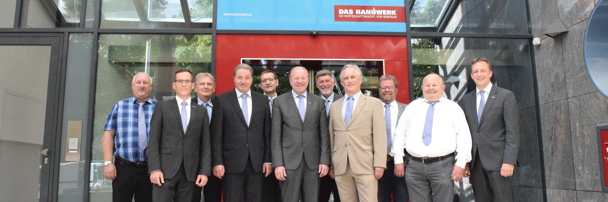Vorstand der Handwerkskammer mit Hauptgeschäftsführer & Stellvertreter Eine Gruppe von elf Männern in Anzug bzw. Hemd und Krawatte steht vor dem Eingang eines Gebäudes und lächelt in die Kamera.