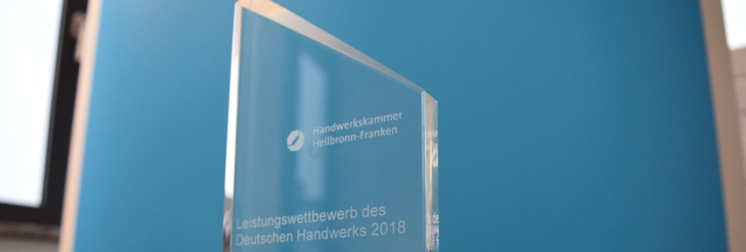Ein gläserner Pokal mit der Aufschrift "Leistungswettbewerb des Deutschen Handwerks 2018" steht vor einem blauen Hintergrund.