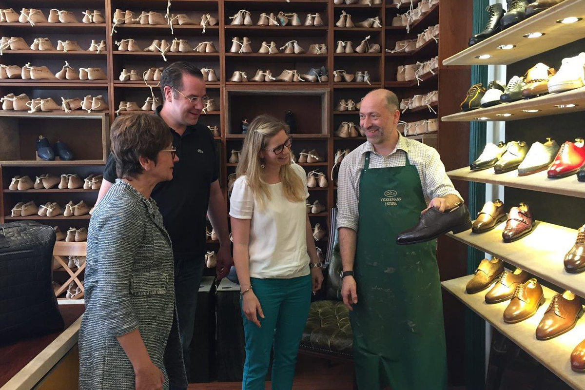 Ein Mann und zwei Frauen stehen in einer Schuhmacher-Werkstatt. Der Schuhmacher zeigt einen Schuh.