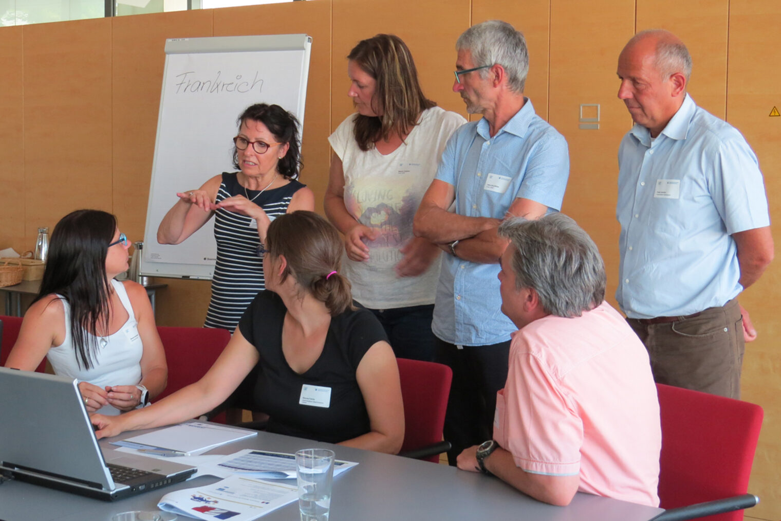 Frauen und Männer diskutieren gemeinsam. Drei davon sitzend an einem Tisch, die anderen vier vor einem Flipchart.