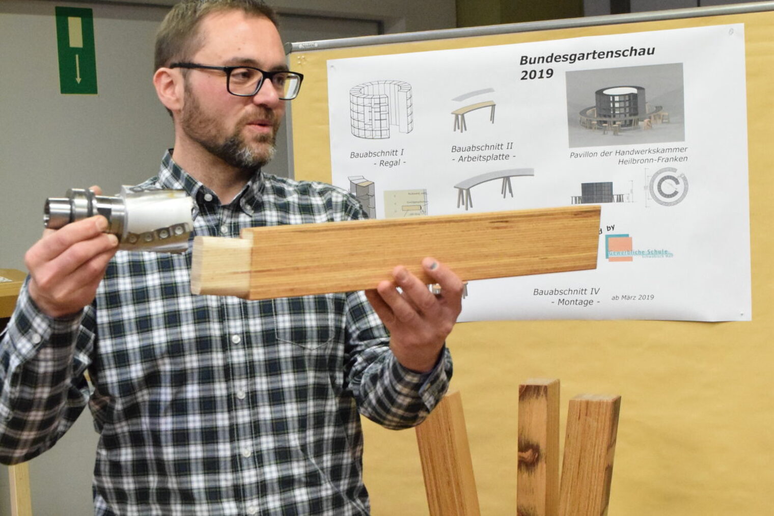 Ein Mann mit Brille und kariertem Hemd hält eine Stück Holz und ein Maschinenteil hoch. Im Hintergrund sieht man Skizzen.