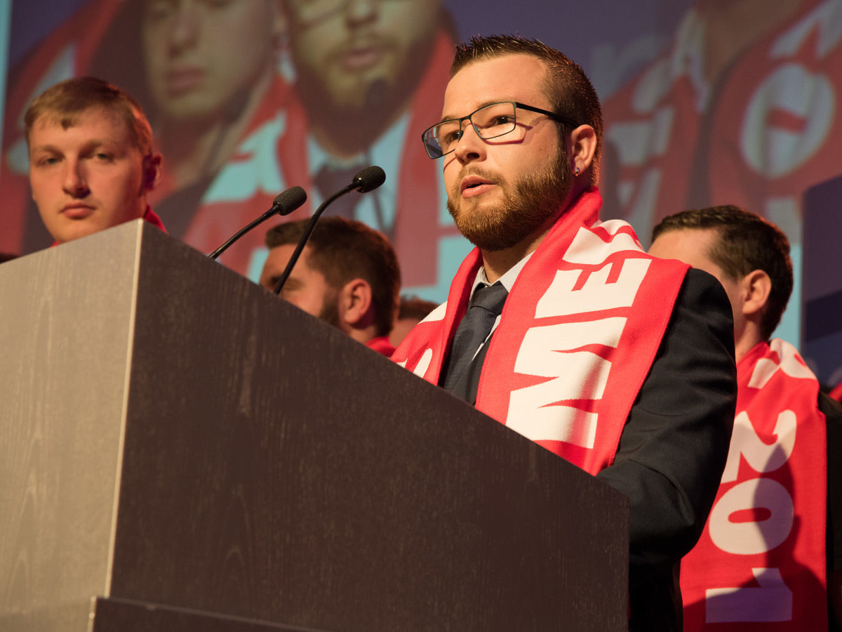 Ein junger Mann mit Brille, Anzug und rotem Schal steht an einem Rednerpult. Im Hintergrund sind weitere Menschen zu sehen.