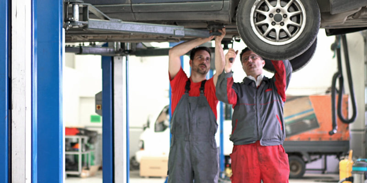 Zwei Männer in Arbeitskleidung stehen in einer Kfz-Werkstatt. Über ihnen sieht man den Teil eines Autos auf einer Hebebühne.