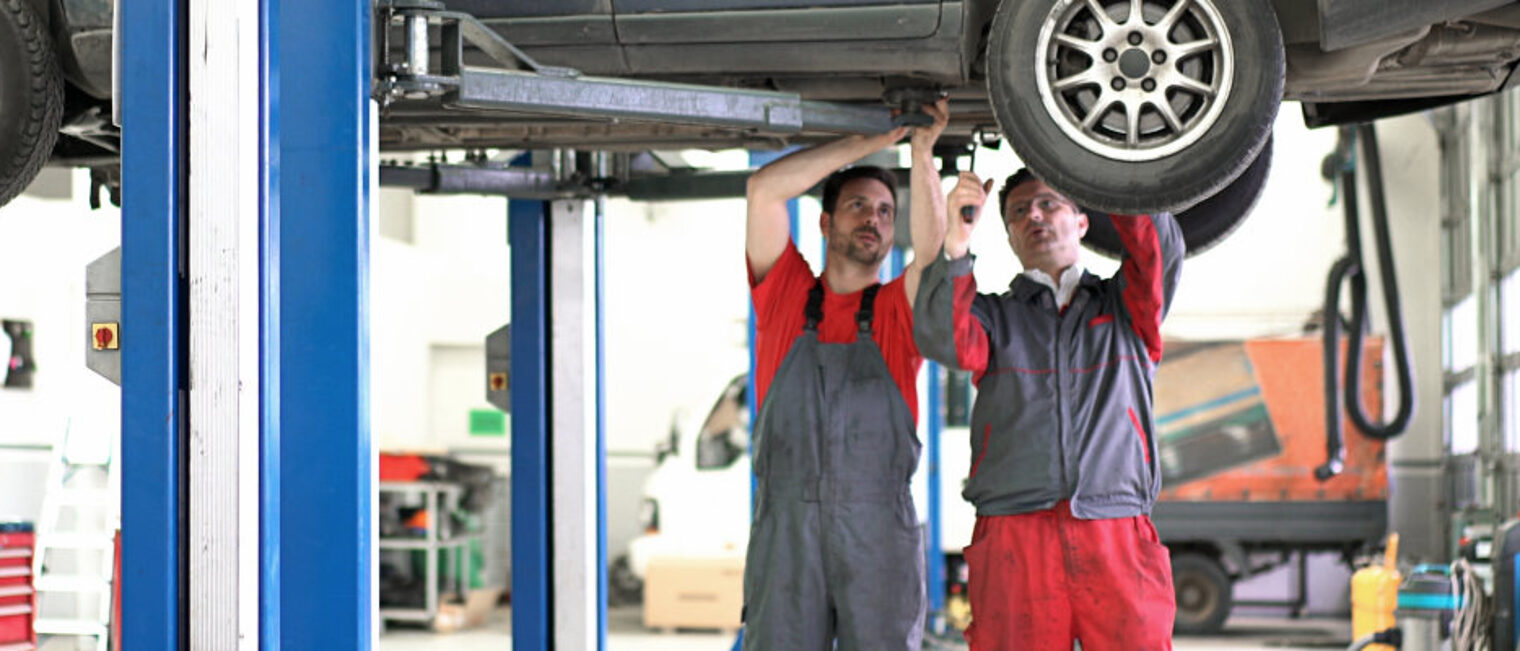 Zwei Männer in Arbeitskleidung stehen in einer Kfz-Werkstatt. Über ihnen sieht man den Teil eines Autos auf einer Hebebühne.