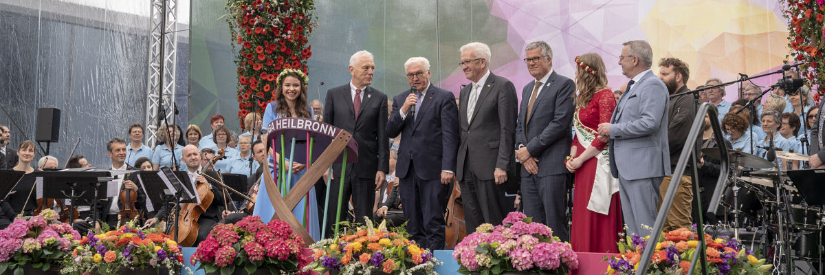 Das Rednerpult der Schreinermeisterschule bei der BUGA-Eröffnung . Eine Gruppe Menschen steht auf einer Bühne. Im Hintergrund ist ein Orchester zu sehen, Im Vordergrund viele Blumen.