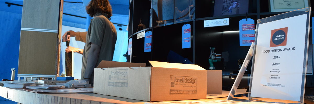 Knelldesign präsentierte im Pavillon sein ausgezeichnetes Regalsystem. Im Vordergrund ein Schild und eine Schachtel mit Aufdruck "knelldesign". Im Hintergrund bauen zwei Männer an einem Regal.