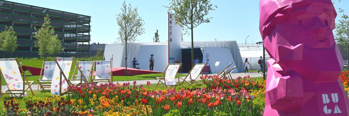 Der Pavillon des Handwerk am Eingang Wohlgelegen der Bundesgartenschau Im Vordergrund ein pinker Gartenzwerg, im Hintergrund eine Blumenwiese, Liegestühle und ein weißer Holzbau.