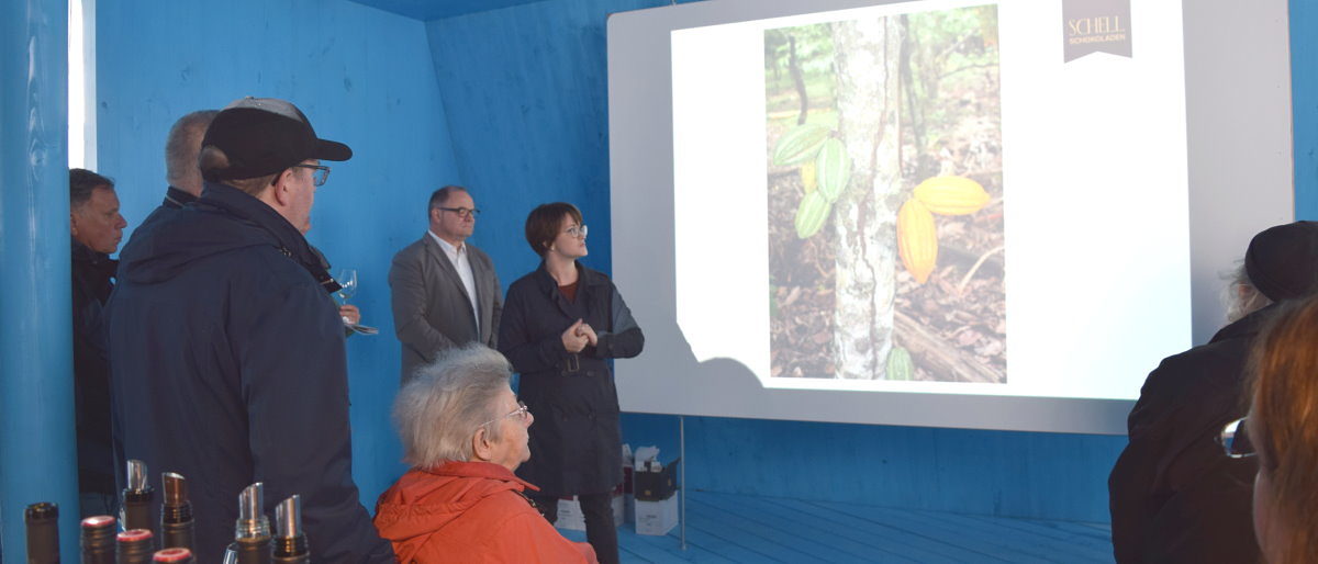 Agnes Reichert-Schell führte durch das Seminar "Schokolade und Wein". Eine Frau steht vor einer Leinwand, auf der ein Foto eines Baums mit einer Frucht zu sehen ist. Mehrere Menschen hören zu.