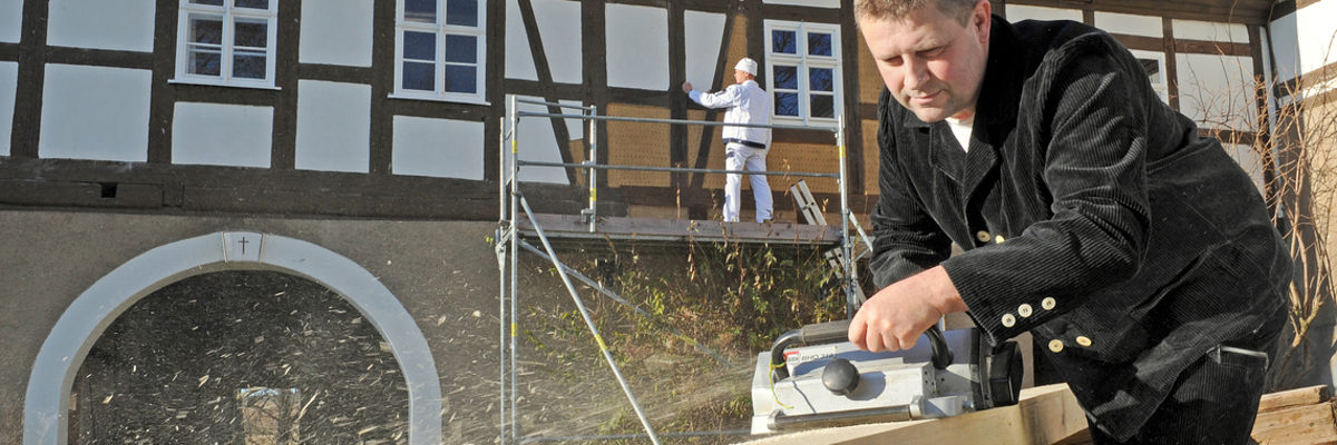 Jetzt für Bundespreis für Handwerk in der Denkmalpflege bewerben. Im Vordergrund schleift ein Mann ein Stück Holz. Im Hintergrund steht ein Mann auf einem Gerüst vor altem Fachwerk.