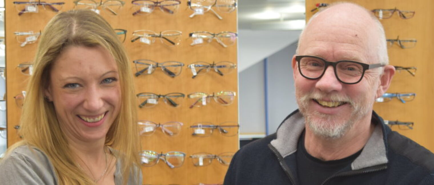 Eine Frau und ein Mann stehen beim Optiker vor einer Wand mit vielen Brillengestellen und lächeln fröhlich in die Kamera.