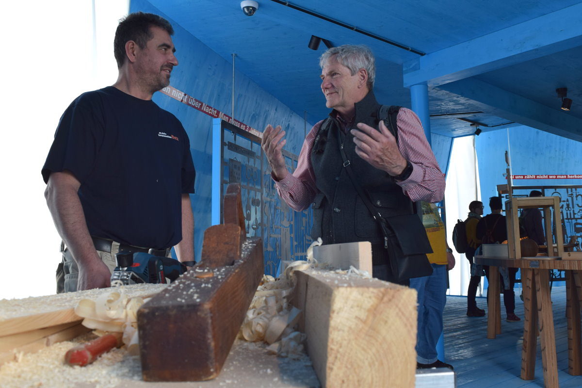 Karl Betz (l.) fachsimpelte mit Besuchern im Pavillon des Handwerks. Zwei Männer sind im Gespräch. Im Vordergrund sind Holzleisten, Hobel und Späne zu sehen.