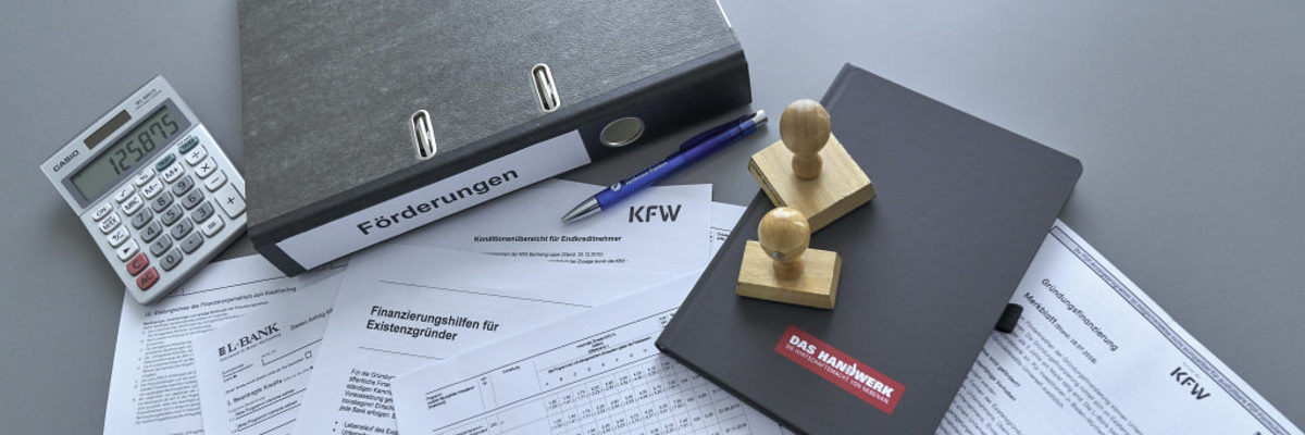 Handwerker erhalten Unterstützung in Sachen Gründungsfinanzierung. Auf einem Tisch liegen verschiedene Antrags- und Merkblätter zu Finanzierung und Fördermöglichkeiten, Ordner, Stempel, Notizbuch