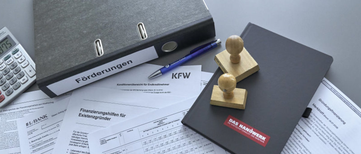 Handwerker erhalten Unterstützung in Sachen Gründungsfinanzierung. Auf einem Tisch liegen verschiedene Antrags- und Merkblätter zu Finanzierung und Fördermöglichkeiten, Ordner, Stempel, Notizbuch