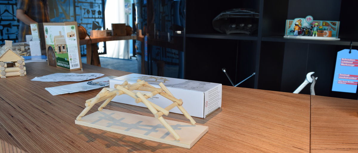 Im Pavillon des Handwerks konnte man kleine Da Vinci-Brücken bauen. Eine Brücke aus runden Holzstäbchen steht auf einem Tisch aus Holz. Im Hintergrund sind weitere Holzkonstruktionen zu sehen.