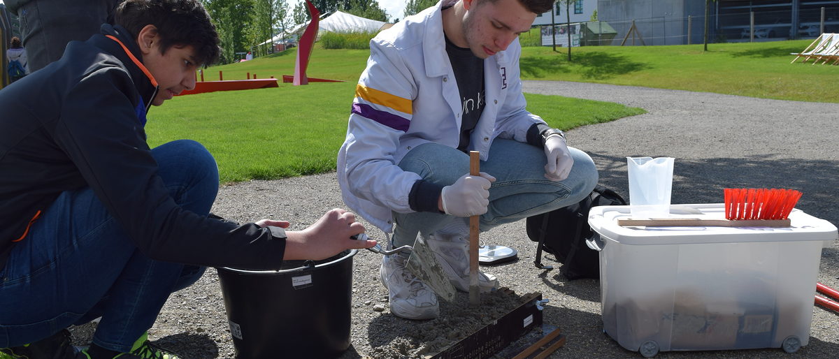 Beim "Bunten Klassenzimmer" können Schüler an mehreren Stationen ausprobieren, wie Handwerk und Architektur zusammenhängen. Zwei Jugendliche sitzen in der Hocke und füllen Beton in eine Holzform.