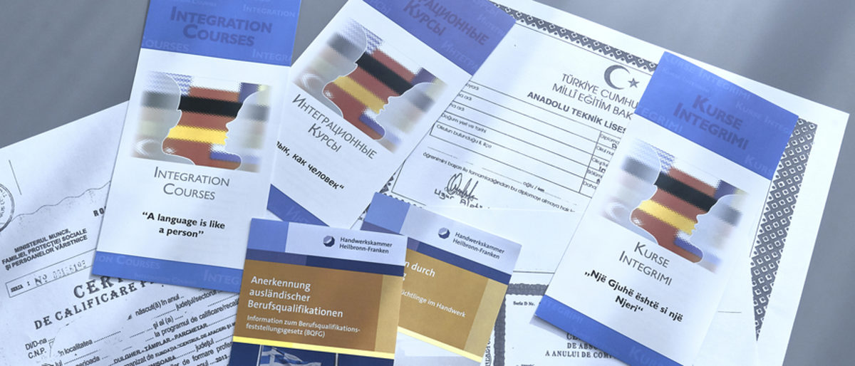 Berufsabschlüsse aus dem Ausland können in Deutschland anerkannt werden. Verschiedene amtliche Dokumente in unterschiedlichen Sprachen und Flyer liegen auf einem grauen Untergrund.