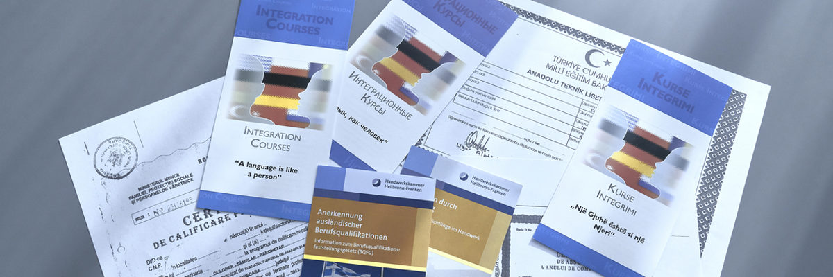 Berufsabschlüsse aus dem Ausland können in Deutschland anerkannt werden. Verschiedene amtliche Dokumente in unterschiedlichen Sprachen und Flyer liegen auf einem grauen Untergrund.