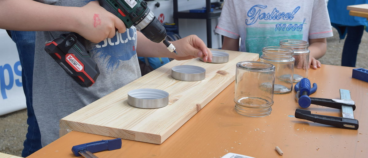 Bei den "Makers-Tagen" können Kinder und Jugendliche Holz- und Gips-Objekte herstellen und Wände gestalten. Ein Holzbrett liegt auf einem Tisch. Daneben sind Schraubzwingen, Gläser und Deckel zu sehen . Kinderhände halten einen Akku-Bohrer in der Hand.