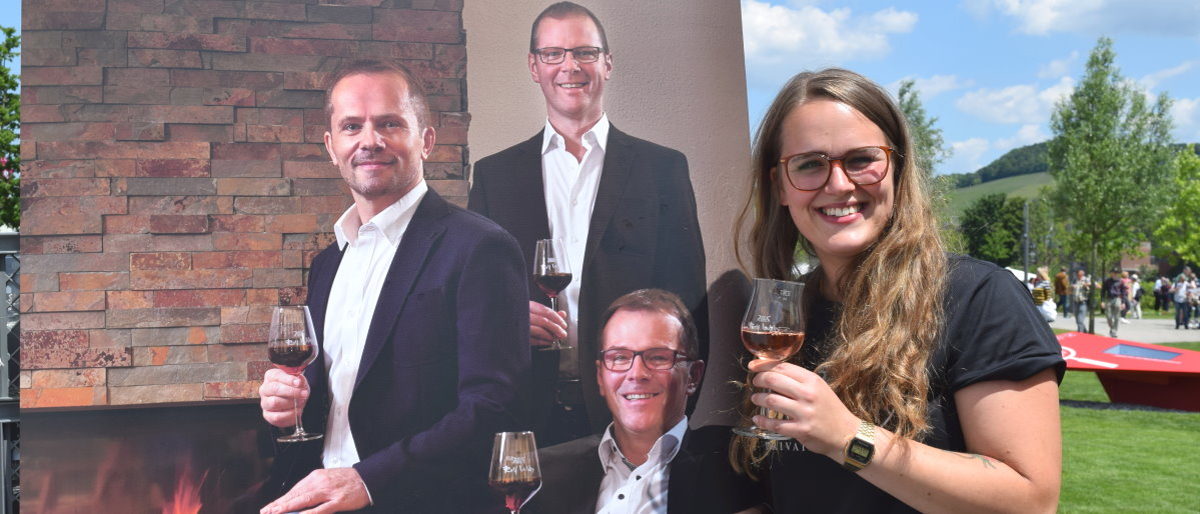 Alina Jans hatte die Idee für die Verkostung von Wein und Brot. Junge Frau hält ein Weinglas in der Hand und lächelt in die Kamera. Neben ihr ein Plakat mit drei Männern mit Weingläsern