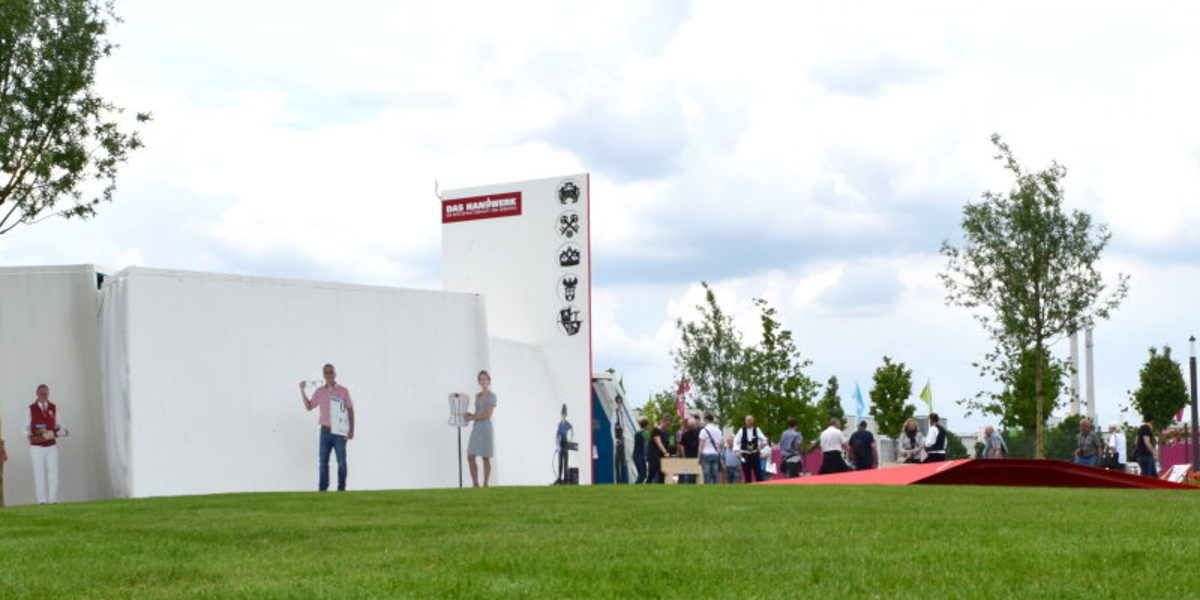 Im Pavillon des Handwerks warten viele Veranstaltungen auf die Besucher Ein weißer Bau steht auf grünem Rasen. Davor stehen Figuren und ein Baum. Rechts steht eine Gruppe Menschen.