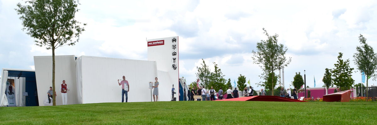 Im Pavillon des Handwerks warten viele Veranstaltungen auf die Besucher Ein weißer Bau steht auf grünem Rasen. Davor stehen Figuren und ein Baum. Rechts steht eine Gruppe Menschen.