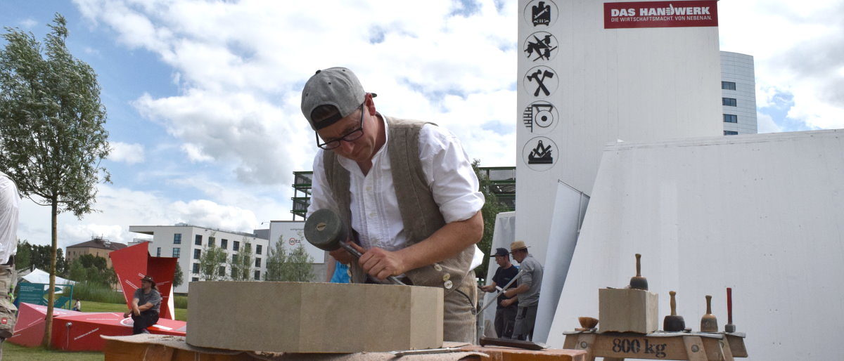 Steinmetzmeister Anton Wachauf bearbeitete auf der BUGA ein Maßwerk. Ein Mann mit Baseball-Kappe und Brille steht mit Werkzeug in der Hand über einen Sandstein gebeugt und bearbeitet ihn.