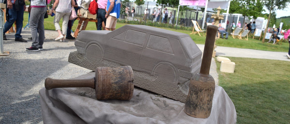 Die Skulptur eines VW Golf zog die Blicke der BUGA-Besucher auf sich. Eine unfertige Skulptur eines Autos aus Stein steht auf einem Sockel. Daneben liegt Werkzeug. Im Hintergrund sind Menschen.