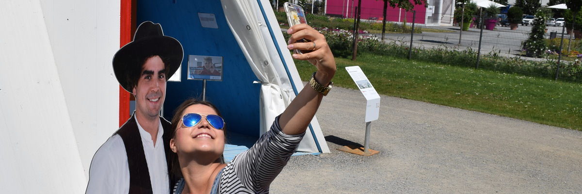 Beim Selfie-Contest der Handwerkskammer ein neues Smartphone gewinnen! Eine junge Frau mit Sonnenbrille hat ein Smartphone in der ausgestreckten Hand. Sie posiert neben der Figur eines Zimmermanns