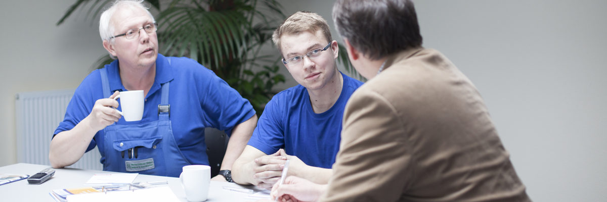 Bei Fragen helfen die Ausbildungsberater Betrieben und Azubis. Drei Männer sitzen am Tisch. Einer im Vordergrund mit dem Rücken zur Kamera, ein junger und ein älterer Mann hören ihm zu.