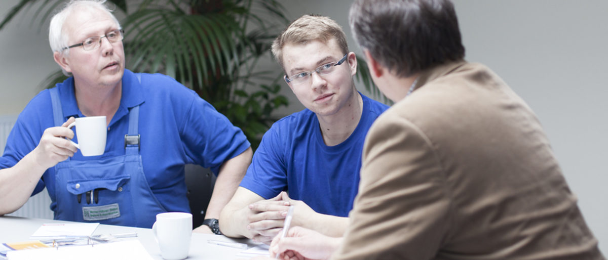 Bei Fragen helfen die Ausbildungsberater Betrieben und Azubis. Drei Männer sitzen am Tisch. Einer im Vordergrund mit dem Rücken zur Kamera, ein junger und ein älterer Mann hören ihm zu.