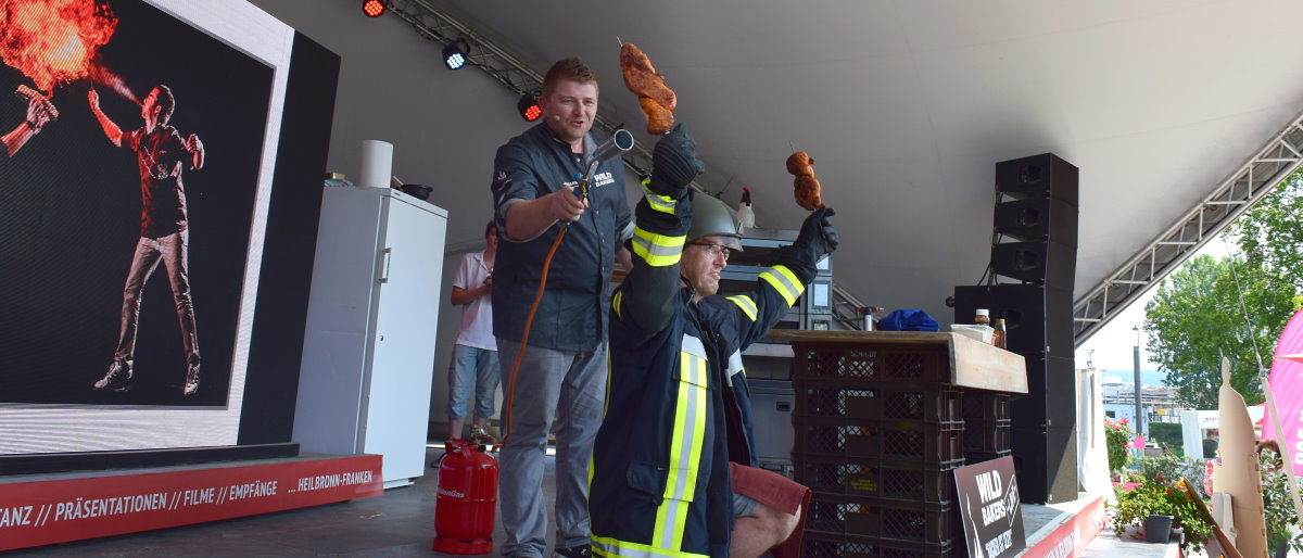 Feuer frei: Ein Wildbaker flambiert sein Brot lieber mit großem Gerät Auf einer Bühne kniet ein Mann in Feuerwehr-Montur. Er hält zwei Brote nach oben, die ein zweiter Mann mit Feuer bearbeitet.