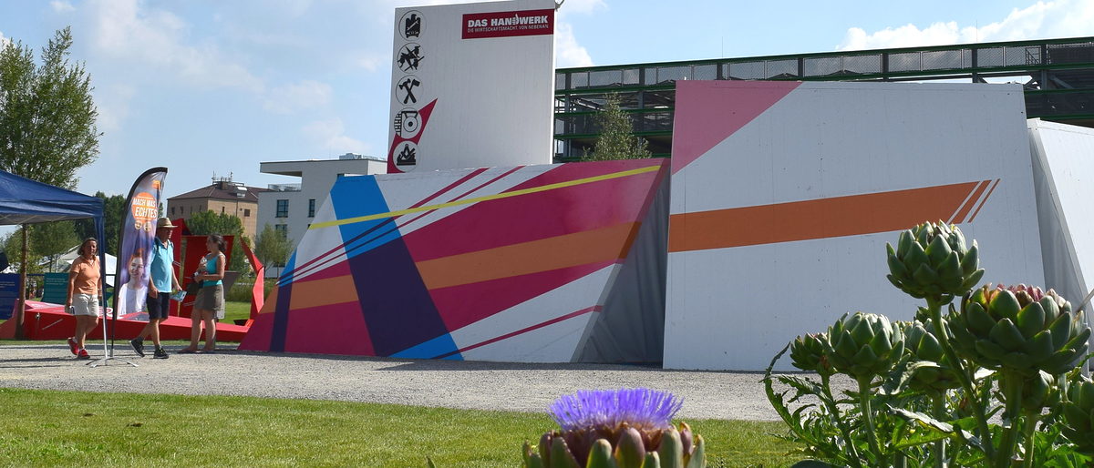 Der Pavillon des Handwerks auf der Bundesgartenschau 2019 in Heilbronn. Eine weiße Holzkonstruktion, mit farbigen Mustern gestaltet, daneben einige Menschen. Im Vordergrund Rasen und Blumen.