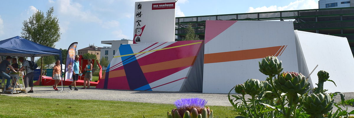 Der Pavillon des Handwerks auf der Bundesgartenschau 2019 in Heilbronn. Eine weiße Holzkonstruktion, mit farbigen Mustern gestaltet, daneben einige Menschen. Im Vordergrund Rasen und Blumen.