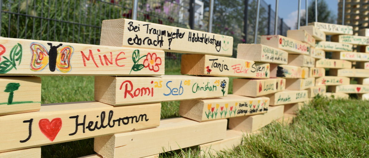 BUGA-Besucher können sich bei der Aktion "The Wall" verewigen. Beschriftete Holzstücke sind auf einer Rasenfläche übereinander geschichtet, so dass eine fortlaufende Wand entsteht.