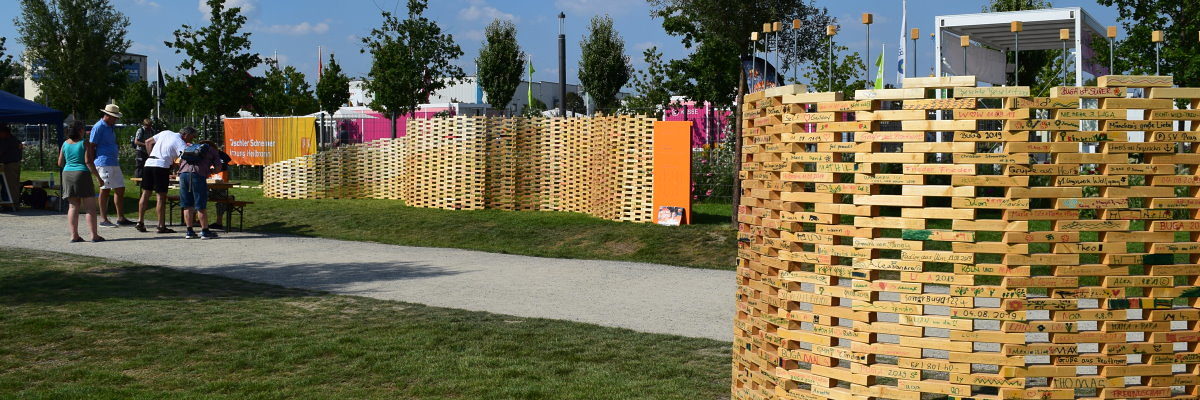 Aktion "The Wall" der Schreiner-Innung Heilbronn auf der BUGA. Zwei geschwungene Wände aus beschrifteten Holzstücken stehen auf zwei Rasenflächen. Dazwischen ein Schotterweg mit Menschen.