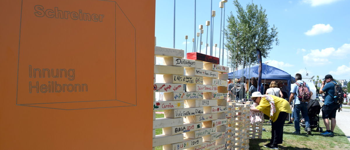 BUGA-Besucher können sich bei der Aktion "The Wall" verewigen. Eine oranges Schild mit Schrift "Schreiner Innung Heilbronn", dahinter eine geschwungene Wand aus beschrifteten Holzstücken.