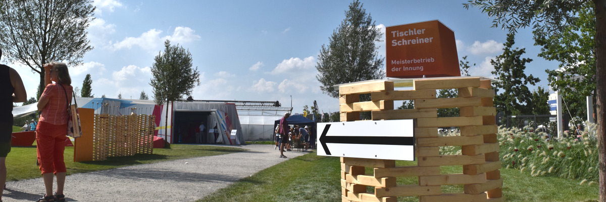 Kommende Veranstaltungen im Pavillon des Handwerks auf der BUGA. Turm aus gestapelten Holzstücken mit einem Pfeil daran, der nach hinten auf einen Holzbau und eine Wand aus Holzklötzen zeigt