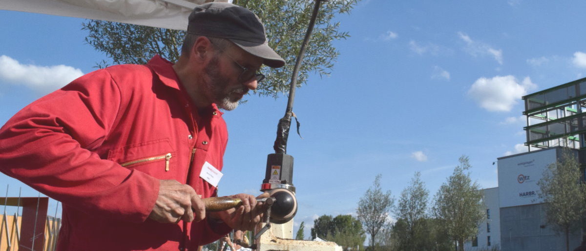 Bernd Pfister fertigte auf der BUGA Holzkugeln an der Drehbank. Ein Mann in rotem Overall und grauer Kappe bearbeitet im Freien ein Stück dunkles Holz an einer Drehbank mit einem Werkzeug.